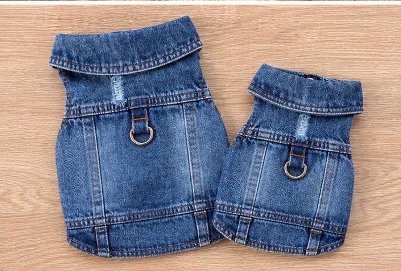 May include: Two blue denim pet vests featuring a fold-down collar, a distressed fabric detail on the upper back, and a silver-toned metal D-ring attachment point centered on the back panel. The vests have visible stitching and a classic denim texture.