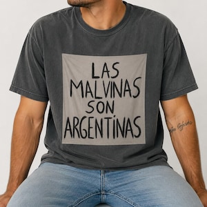 May include: A charcoal gray short-sleeve t-shirt featuring a large, centered, light gray square patch. The patch displays the black hand-lettered text "LAS MALVINAS SON ARGENTINAS" in a bold, informal font style across three lines.