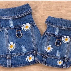 May include: Two blue denim vests featuring a distressed finish, a fold-down collar, and white daisy flower embroidery with yellow centers. Each vest includes a metal D-ring attachment on the back for harness functionality and visible stitching.