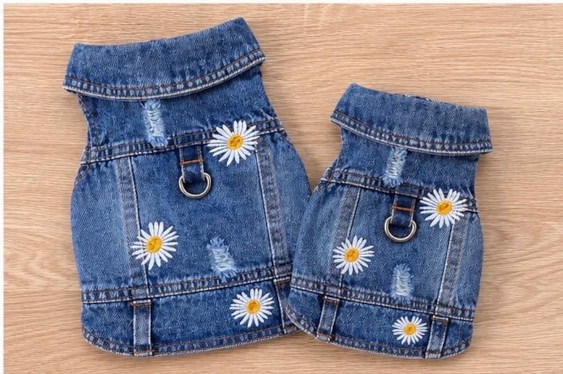 May include: Two blue denim vests featuring a distressed finish, a fold-down collar, and white daisy flower embroidery with yellow centers. Each vest includes a metal D-ring attachment on the back for harness functionality and visible stitching.