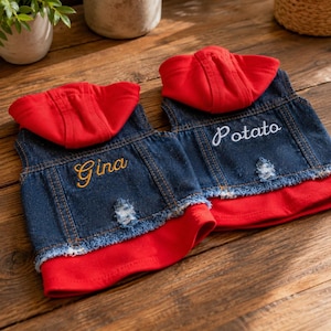 May include: Two denim vests with red hoods and red undershirts. The left vest features the embroidered name Gina in yellow, and the right vest features the embroidered name Potato in white. Both vests have distressed frayed hems.