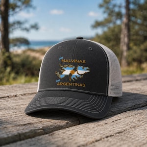 May include: A charcoal gray trucker hat with a mesh back and a curved brim. The front panel features a graphic of the Falkland Islands filled with the Argentine flag colors and a sun emblem, with the text "las MALVINAS son ARGENTINAS" in yellow.