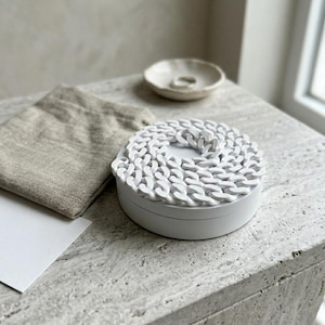 Könnte beinhalten: Eine weiße, runde Schmuckschatulle mit einem Kettendesign auf dem Deckel. Eine kleine, weiße Keramikschale und ein Leinenbeutel befinden sich neben der Schatulle. Die Artikel stehen auf einer hellen, strukturierten Oberfläche.