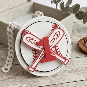 May include: A white and red baseball-themed ornament. The design features a baseball with crossed bats and the number 1 in red. The ornament is round with a black border and a white chain.