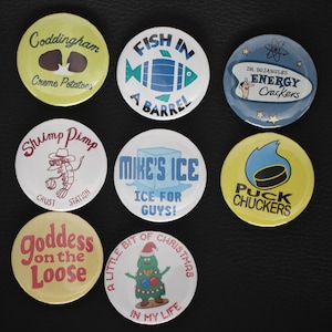 May include: A collection of eight round pinback buttons with various designs and text. The buttons feature phrases like "Fish in a Barrel," "Shrimp Pimp," and "Puck Chuckers." Colors include yellow, blue, white, and red.