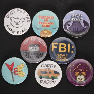 May include: Eight circular pin badges with diverse designs and text. The badges display illustrations of a bear, pancakes, a mountain scene, a cartoon character, the letters "FBI", a guitar, a camper van, and a clock.
