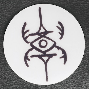 May include: A white, circular sticker with a dark purple, stylized sigil. The design incorporates an eye shape, a vertical line, and curved elements that resemble antlers or horns. The sticker is on a black background.