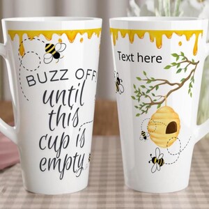 May include: White ceramic latte mug with a yellow honey-dripping design at the top. One side features the text "Buzz Off until this cup is empty" with bee illustrations. The other side has a beehive and the text "Text here".