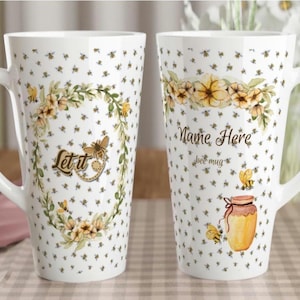 May include: Two white ceramic mugs with bee-themed designs. One mug features a floral wreath with a butterfly and the words "Let it". The other mug has a floral design, the words "Name Here", and a honey pot illustration.