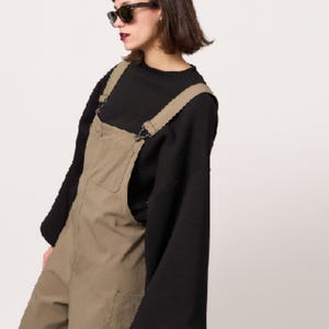 May include: Olive green dungarees featuring a front chest pocket and adjustable shoulder straps with metal hardware, worn over a black long-sleeved top with wide sleeves and a mock neck collar. The dungarees have a textured fabric finish.