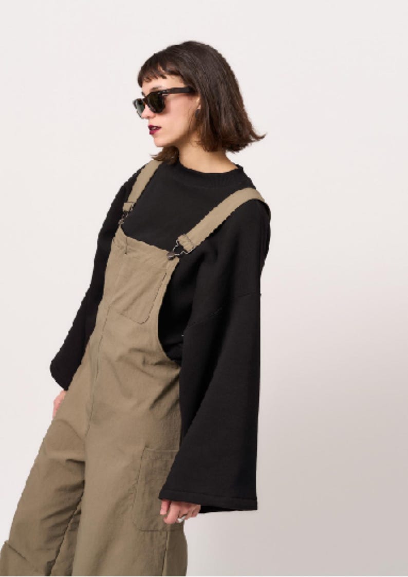 May include: Olive green dungarees featuring a front chest pocket and adjustable shoulder straps with metal hardware, worn over a black long-sleeved top with wide sleeves and a mock neck collar. The dungarees have a textured fabric finish.