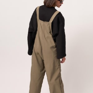 May include: Olive green dungarees featuring wide shoulder straps, a high back, and loose-fitting legs. The garment is worn over a black long-sleeved shirt with a mock neck collar. The dungarees have visible side pockets and a straight-leg silhouette.