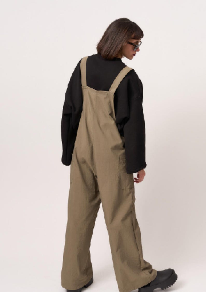 May include: Olive green dungarees featuring wide shoulder straps, a high back, and loose-fitting legs. The garment is worn over a black long-sleeved shirt with a mock neck collar. The dungarees have visible side pockets and a straight-leg silhouette.