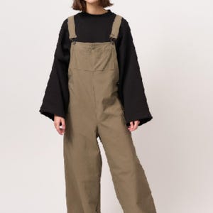 May include: Olive green cotton dungarees featuring a large front bib pocket, adjustable shoulder straps with metal buckles, and wide-leg trousers. The dungarees are worn over a black long-sleeved top with wide sleeves and paired with black platform shoes.
