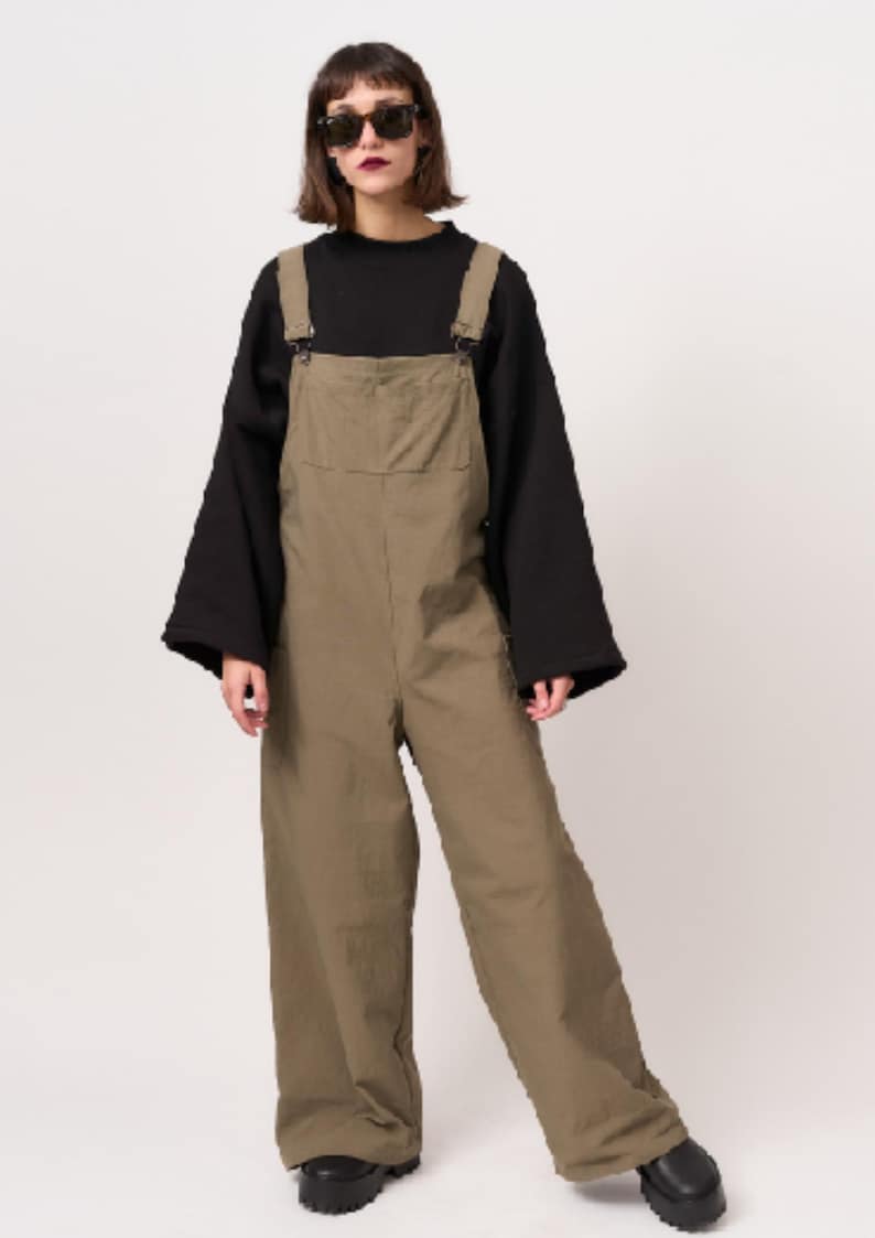 May include: Olive green cotton dungarees featuring a large front bib pocket, adjustable shoulder straps with metal buckles, and wide-leg trousers. The dungarees are worn over a black long-sleeved top with wide sleeves and paired with black platform shoes.