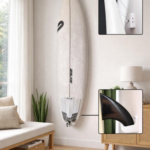 Könnte beinhalten: Ein weißes Surfbrett-Wandhalterungssystem mit einer oberen Drahtseilhalterung und einer unteren Finnenhalterung. Das Surfbrett ist weiß mit schwarzem Schriftzug und einem strukturierten weißen Traktionspad. Die Halterungen sind weiß.