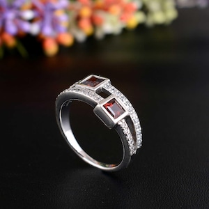 Könnte beinhalten: Ein silberner Ring mit zwei quadratischen, roten Edelsteinen und einem Band, das mit kleinen, klaren Steinen besetzt ist. Der Ring ist vor einem schwarzen Hintergrund mit verschwommenen Blumen im Hintergrund platziert.