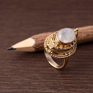 Peut inclure: Une bague dorée ornée d'une pierre précieuse ovale et claire. La bague présente des détails complexes autour de la pierre et sur le corps. Elle est posée sur une surface en bois sombre avec un crayon en arrière-plan. Bijoux.