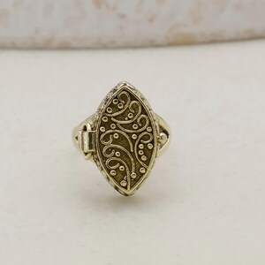 May include: A gold-coloured ring with an oval-shaped face. The face features an ornate, swirling design with small dot accents. The ring has a hinged clasp on the side and a simple band.