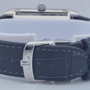 Pu&ograve; includere: Primo piano di un orologio di lusso con cinturino in pelle blu scuro. L'orologio presenta un quadrante rettangolare color argento e una fibbia abbinata. Il cinturino &egrave; cucito con filo blu chiaro.