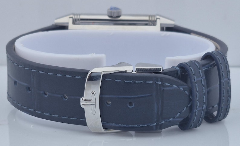 Pu&ograve; includere: Primo piano di un orologio di lusso con cinturino in pelle blu scuro. L'orologio presenta un quadrante rettangolare color argento e una fibbia abbinata. Il cinturino &egrave; cucito con filo blu chiaro.
