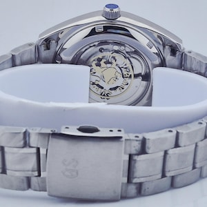 May include: A silver-toned wristwatch with a visible mechanical movement and a stainless steel band. The watch features a blue crown and a clasp with a logo. The watch is displayed on a white support.
