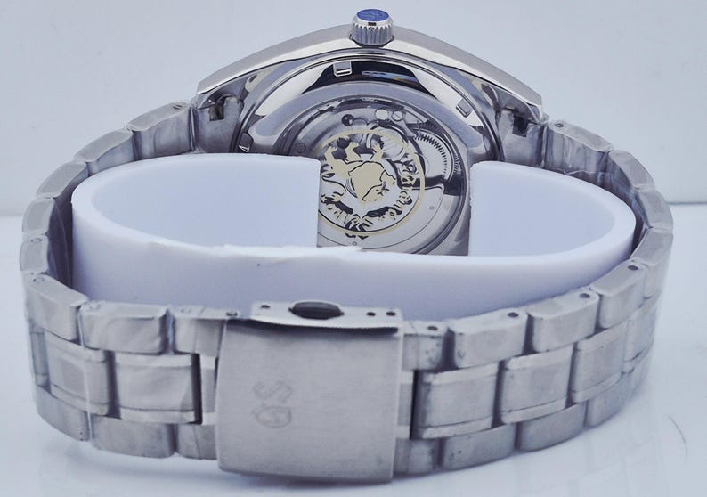May include: A silver-toned wristwatch with a visible mechanical movement and a stainless steel band. The watch features a blue crown and a clasp with a logo. The watch is displayed on a white support.