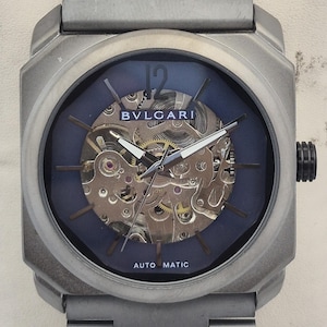 May include: A silver-toned Bulgari automatic watch with a blue face and visible internal gears. The watch has a rectangular case and a matching metal band. The watch face displays the brand name and the words "AUTO MATIC".