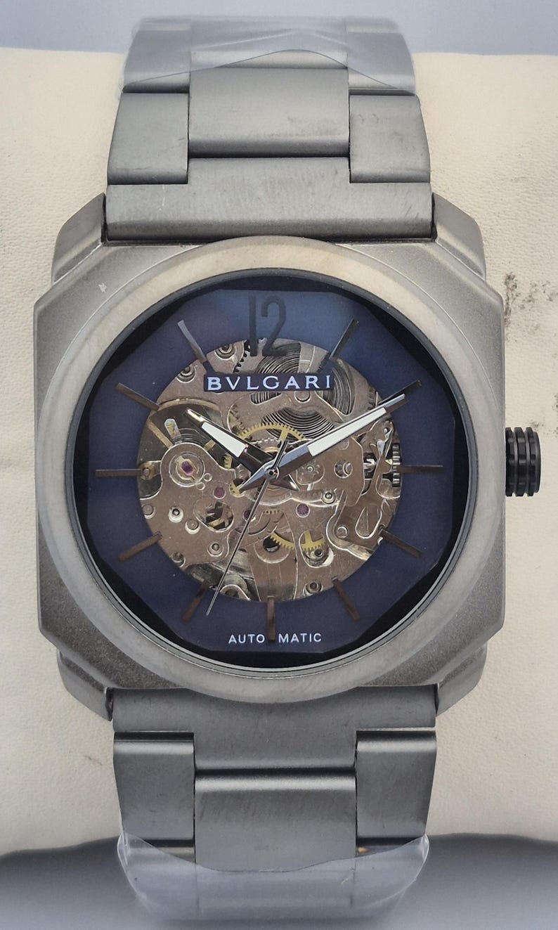 May include: A silver-toned Bulgari automatic watch with a blue face and visible internal gears. The watch has a rectangular case and a matching metal band. The watch face displays the brand name and the words "AUTO MATIC".