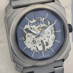 May include: A silver-toned wristwatch with a blue face and visible internal mechanisms. The watch features the brand name "BVLGARI" and the word "AUTOMATIC". The watch has a silver-toned band and a black crown.