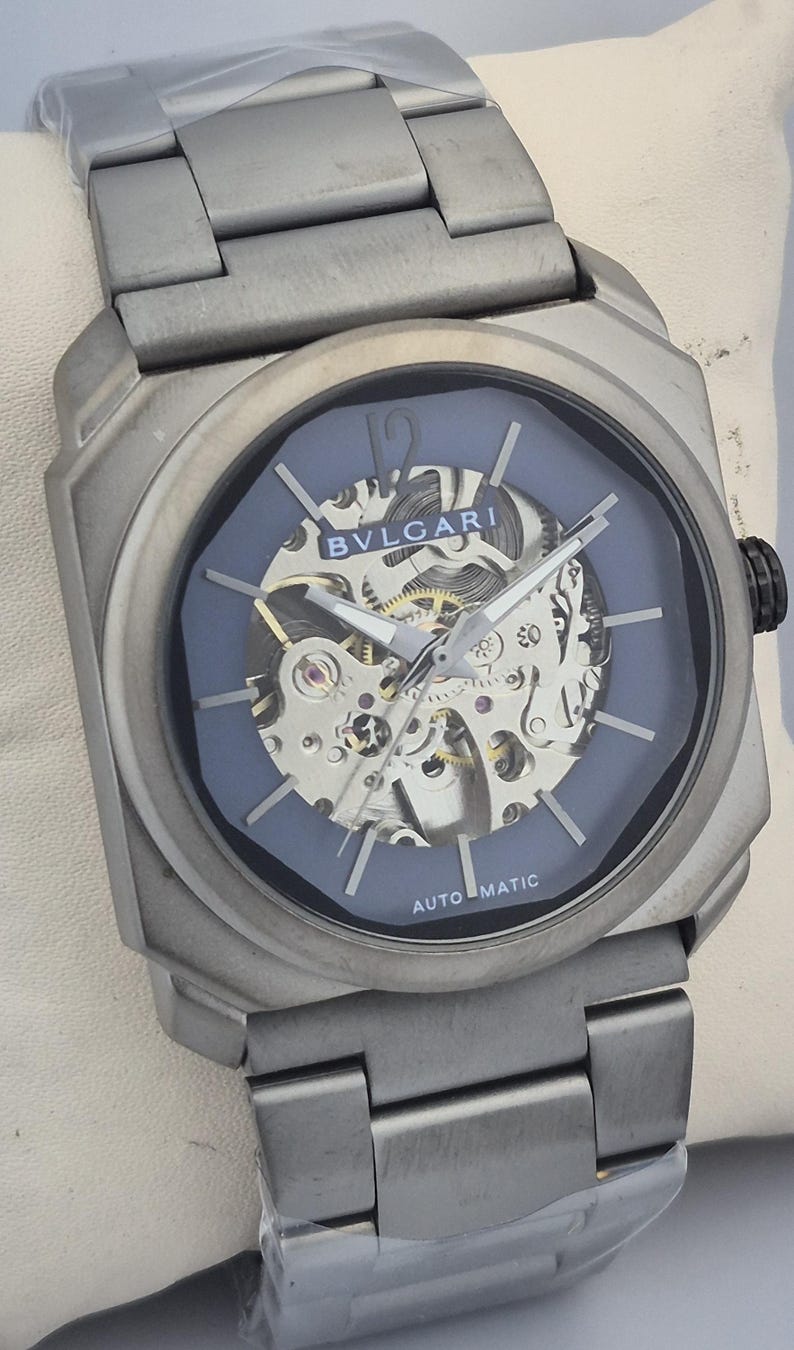 May include: A silver-toned wristwatch with a blue face and visible internal mechanisms. The watch features the brand name "BVLGARI" and the word "AUTOMATIC". The watch has a silver-toned band and a black crown.