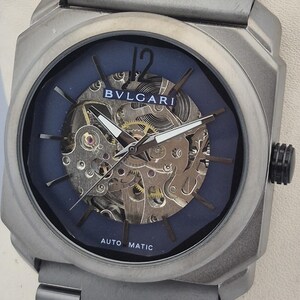 May include: A silver-toned wristwatch with a blue face and visible internal mechanisms. The watch features the brand name "BVLGARI" and the words "AUTO MATIC". The watch has a silver band and a black crown.