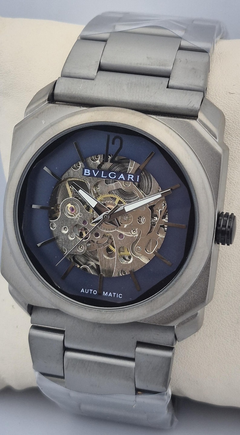 May include: A silver-toned wristwatch with a blue face and visible internal mechanisms. The watch features the brand name "BVLGARI" and the words "AUTO MATIC". The watch has a silver band and a black crown.