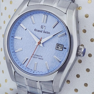 May include: A silver-toned Grand Seiko automatic wristwatch with a light blue dial. The watch features a date display, silver hands, and a red seconds hand. The watch band is also silver-toned.