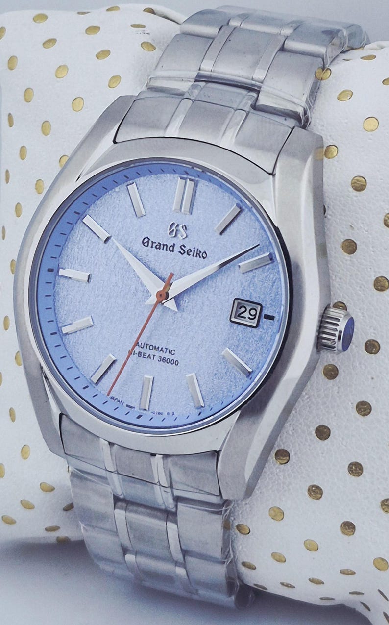 May include: A silver-toned Grand Seiko automatic wristwatch with a light blue dial. The watch features a date display, silver hands, and a red seconds hand. The watch band is also silver-toned.
