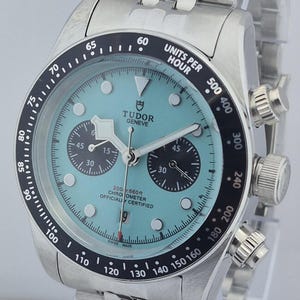 May include: A stainless steel wristwatch with a light blue face and black bezel. The watch features multiple sub-dials, a date display, and the text "TUDOR GENEVE". The watch band is made of stainless steel.
