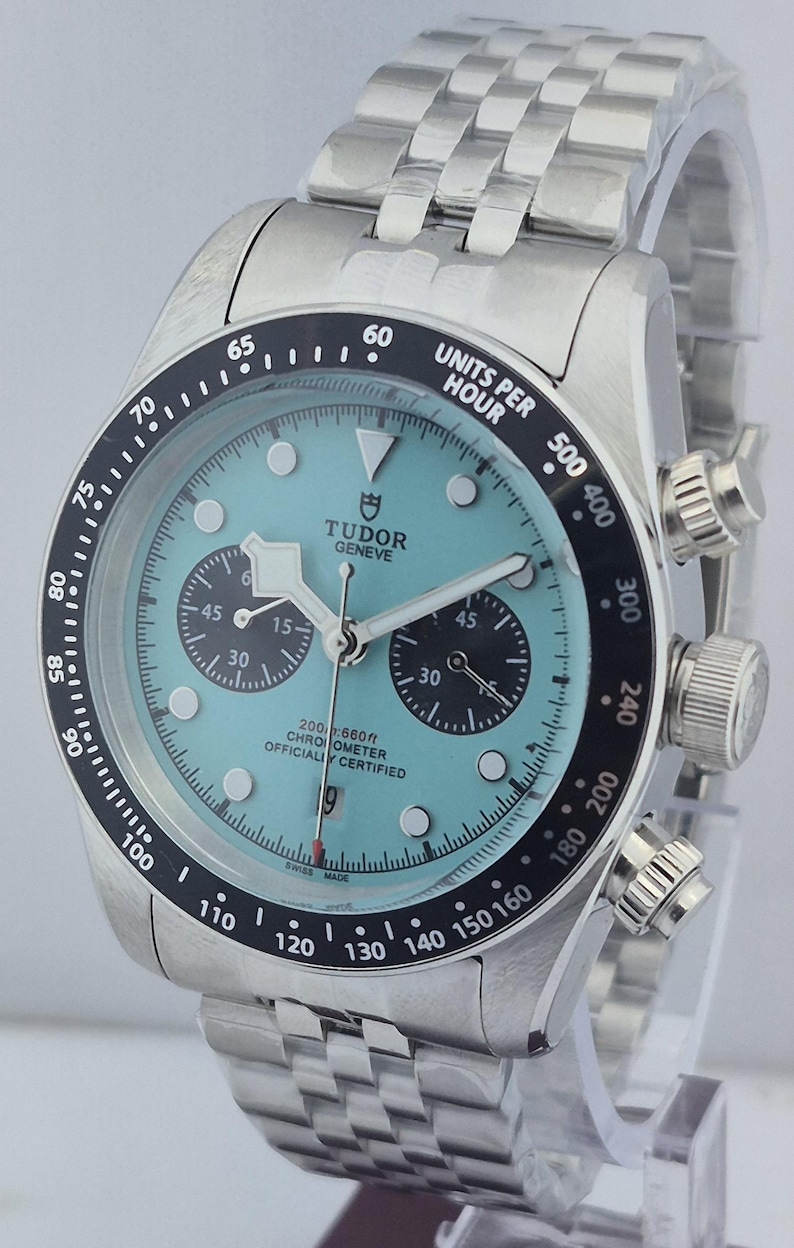May include: A stainless steel wristwatch with a light blue face and black bezel. The watch features multiple sub-dials, a date display, and the text "TUDOR GENEVE". The watch band is made of stainless steel.