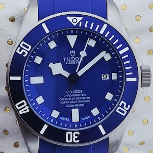 May include: A blue and silver wristwatch with a blue band. The watch face is blue with white markers and hands. The watch has the text "TUDOR GENEVE PELAGOS CHRONOMETER" and "500m-1640ft". The watch is on a white surface with gold dots.