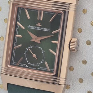 May include: A luxury wristwatch with a rectangular rose gold case and a green leather strap. The watch face is green with rose gold hands and markers, and features the Jaeger-LeCoultre logo. The watch has a small seconds dial and the words "SWISS MADE".