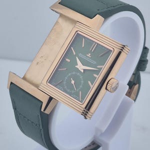 May include: A luxury wristwatch with a rectangular green face and gold-toned case. The watch features a small seconds dial and is set on a green leather strap. The watch is displayed on a white stand.