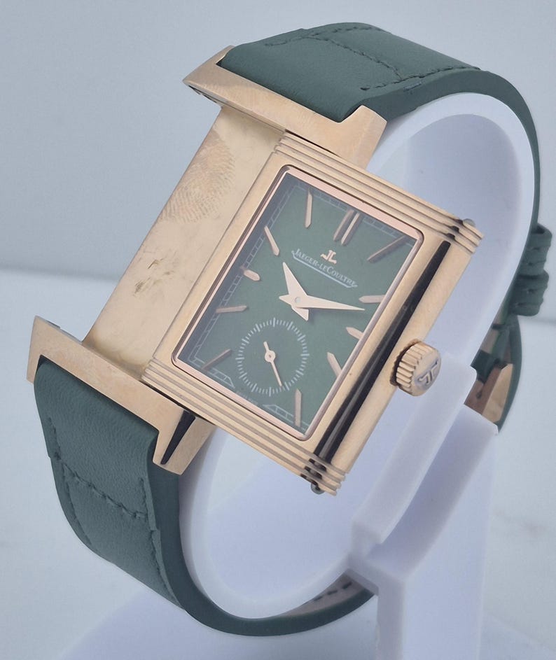 May include: A luxury wristwatch with a rectangular green face and gold-toned case. The watch features a small seconds dial and is set on a green leather strap. The watch is displayed on a white stand.