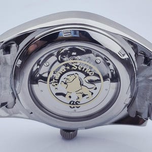 May include: Close-up of a stainless steel wristwatch. The watch's back reveals the intricate inner workings, including gears and a gold-toned emblem with a lion. The watch band is also stainless steel.