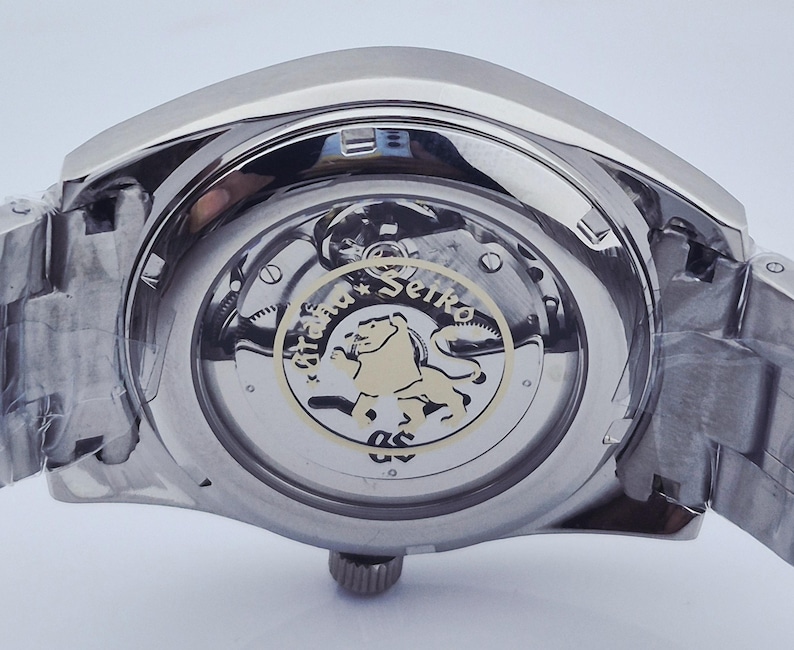 May include: Close-up of a stainless steel wristwatch. The watch's back reveals the intricate inner workings, including gears and a gold-toned emblem with a lion. The watch band is also stainless steel.