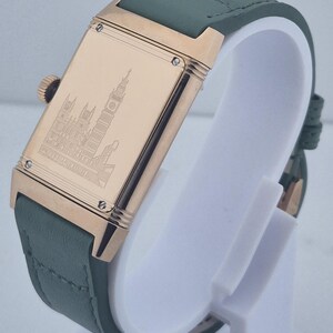 May include: A rectangular wristwatch with a gold-colored case and a green leather strap. The watch face features a detailed engraving of a building. The watch is displayed on a white stand.