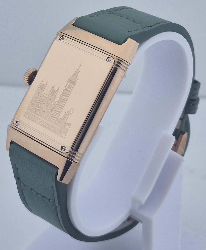 May include: A rectangular wristwatch with a gold-colored case and a green leather strap. The watch face features a detailed engraving of a building. The watch is displayed on a white stand.