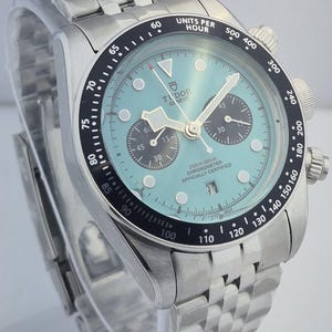 May include: A stainless steel wristwatch with a light blue face and a black bezel. The watch features a chronograph function with three sub-dials and a date display. The watch has a stainless steel bracelet and is water-resistant to 200 meters.