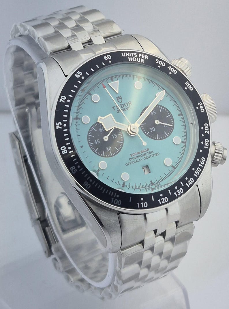May include: A stainless steel wristwatch with a light blue face and a black bezel. The watch features a chronograph function with three sub-dials and a date display. The watch has a stainless steel bracelet and is water-resistant to 200 meters.