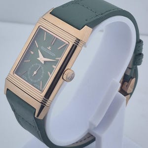 May include: A rectangular wristwatch with a green face and a small seconds dial. The watch has a gold-colored case and hands, and a green leather strap. The watch is displayed on a white stand.