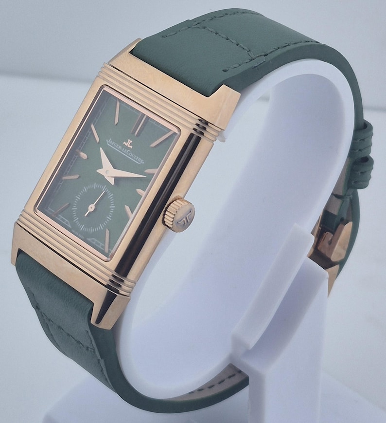 May include: A rectangular wristwatch with a green face and a small seconds dial. The watch has a gold-colored case and hands, and a green leather strap. The watch is displayed on a white stand.