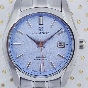 May include: A silver-toned Grand Seiko automatic wristwatch with a light blue dial. The watch features a date display, silver hands, and a silver bracelet. The dial has the text "Grand Seiko" and "Automatic Hi-Beat 36000".